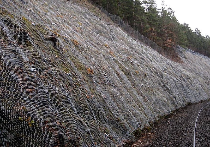Rock slope stabilization system with high-tensile wire mesh drapery and rock bolt anchors protecting an infrastructure corridor